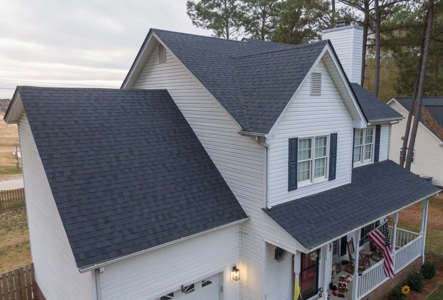 A two-story white house with a dark gray shingle roof, recently serviced by expert Delaware remodeling contractors, is shown. It features a front porch with a small American flag and a cozy seating area. The home is framed by tall trees against an overcast sky.