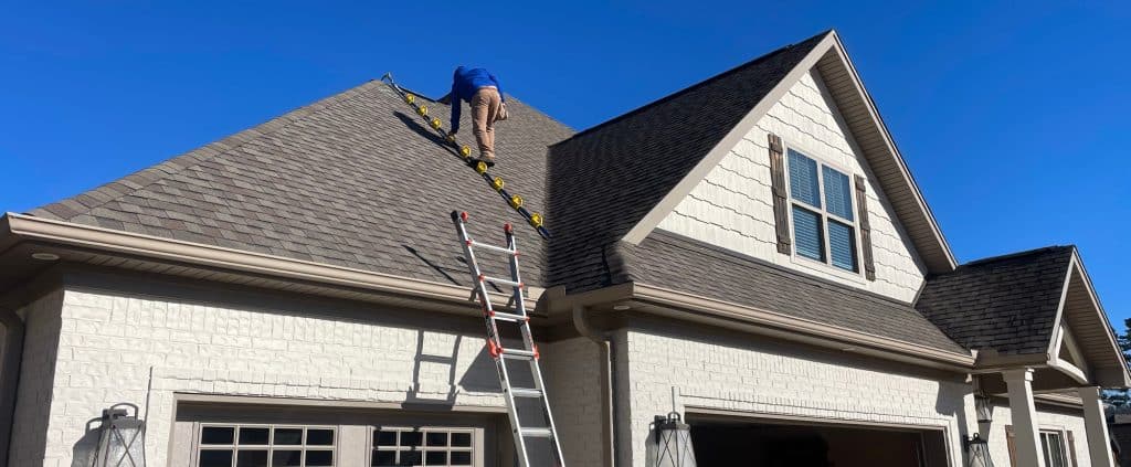 A person in a blue shirt is climbing a ladder to repair the roof. The steep, shingled surface of the Delaware home contrasts with its tan siding and windows, all under a clear blue sky.