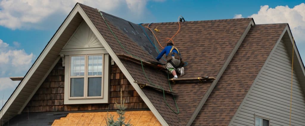 A worker in a blue shirt is expertly handling roof repair on the brown shingle rooftop of a two-story house in Delaware. Surrounded by tools and safety harnesses, he ensures every detail is perfect. The sky above is clear with a few clouds, ideal for such intricate work.