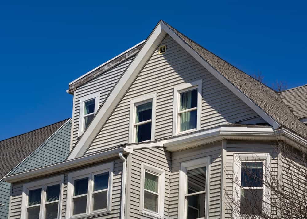 A close-up view of the upper floors of a house with light gray vinyl siding and multiple windows against a clear blue sky. The roof has a gabled design, with one section covered in darker gray shingles.
