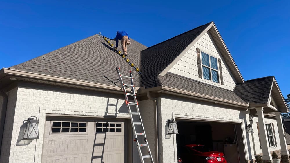 A person climbs a yellow ladder on the steep roof of a light-colored house with two garage doors and a red car inside, under a clear blue sky. Another ladder leans against the garage, suggesting ongoing remodeling services.